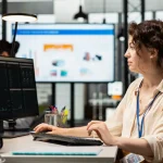 Woman with a blue lanyard typing at a workstation with dual monitors in a modern office environment.