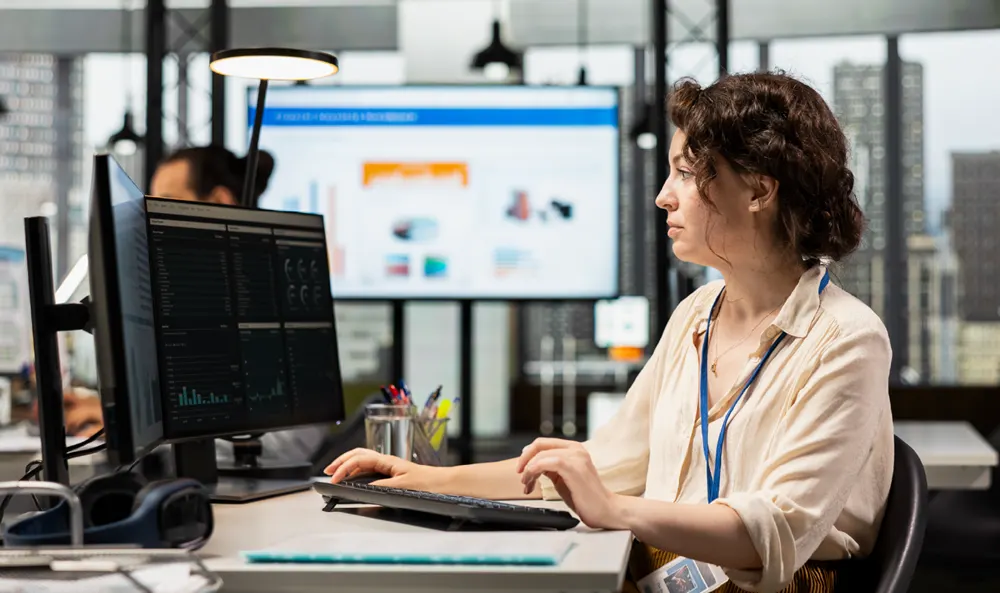 Woman with a blue lanyard typing at a workstation with dual monitors in a modern office environment.